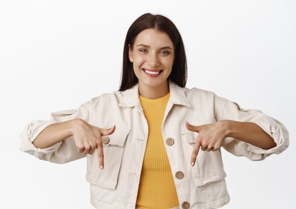 Image of smiling candid woman, pleasant friendly face expression, pointing fingers down, showing sale advertisement below, standing over white background. Copy space